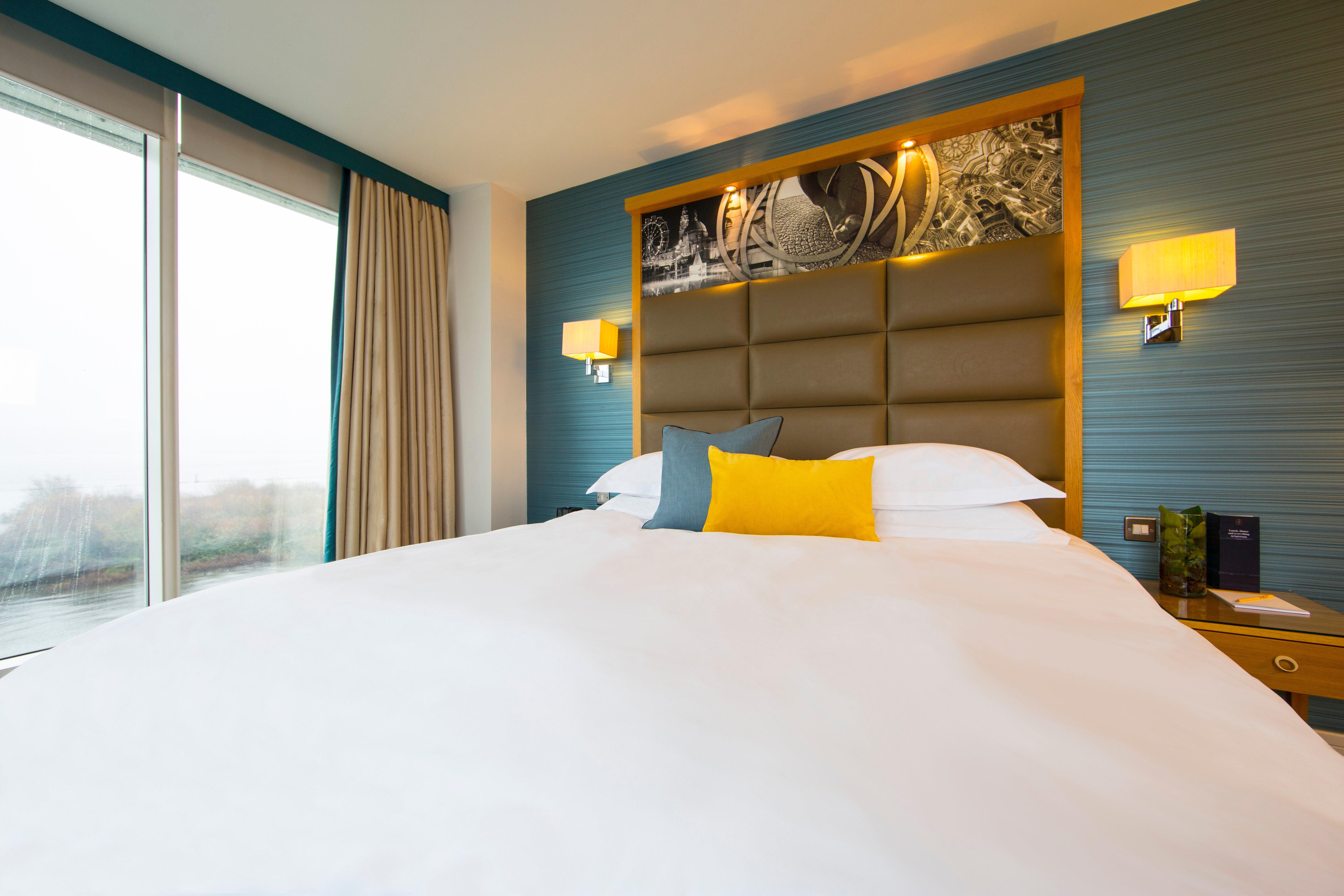فندق فوكو سانت دايفيدز كارديف المظهر الخارجي الصورة The photo shows a modern hotel room featuring a large bed with a neatly made white duvet. The headboard is upholstered in a soft gray material, complemented by decorative lighting on either side. Above the headboard, there is a stylish wall art displ