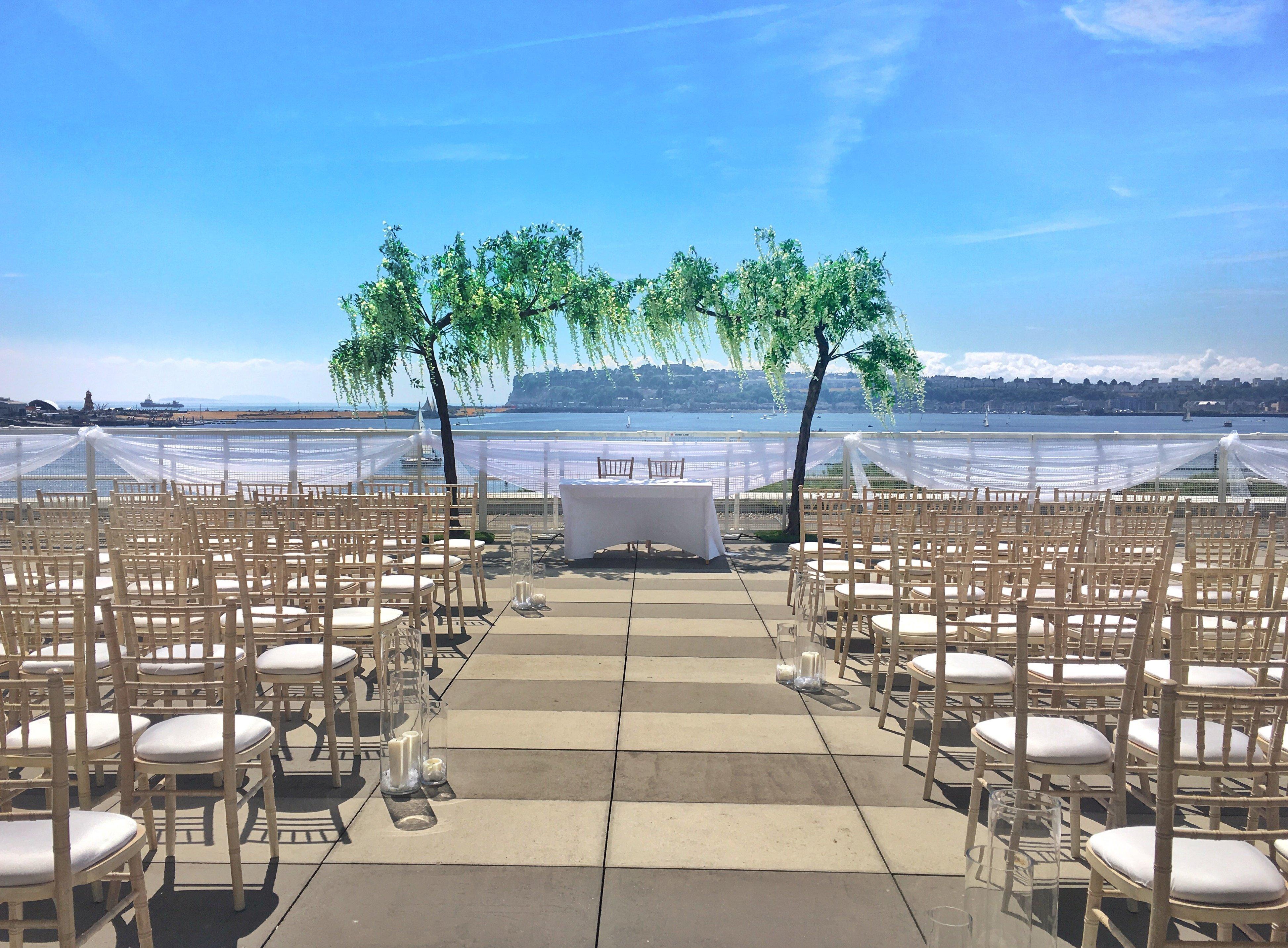 فندق فوكو سانت دايفيدز كارديف المظهر الخارجي الصورة The photo shows a beautiful outdoor wedding venue set up near a body of water. There are rows of wooden chairs arranged in front of a centrally placed white table, likely meant for the wedding ceremony. On either side of the table, there are decorati