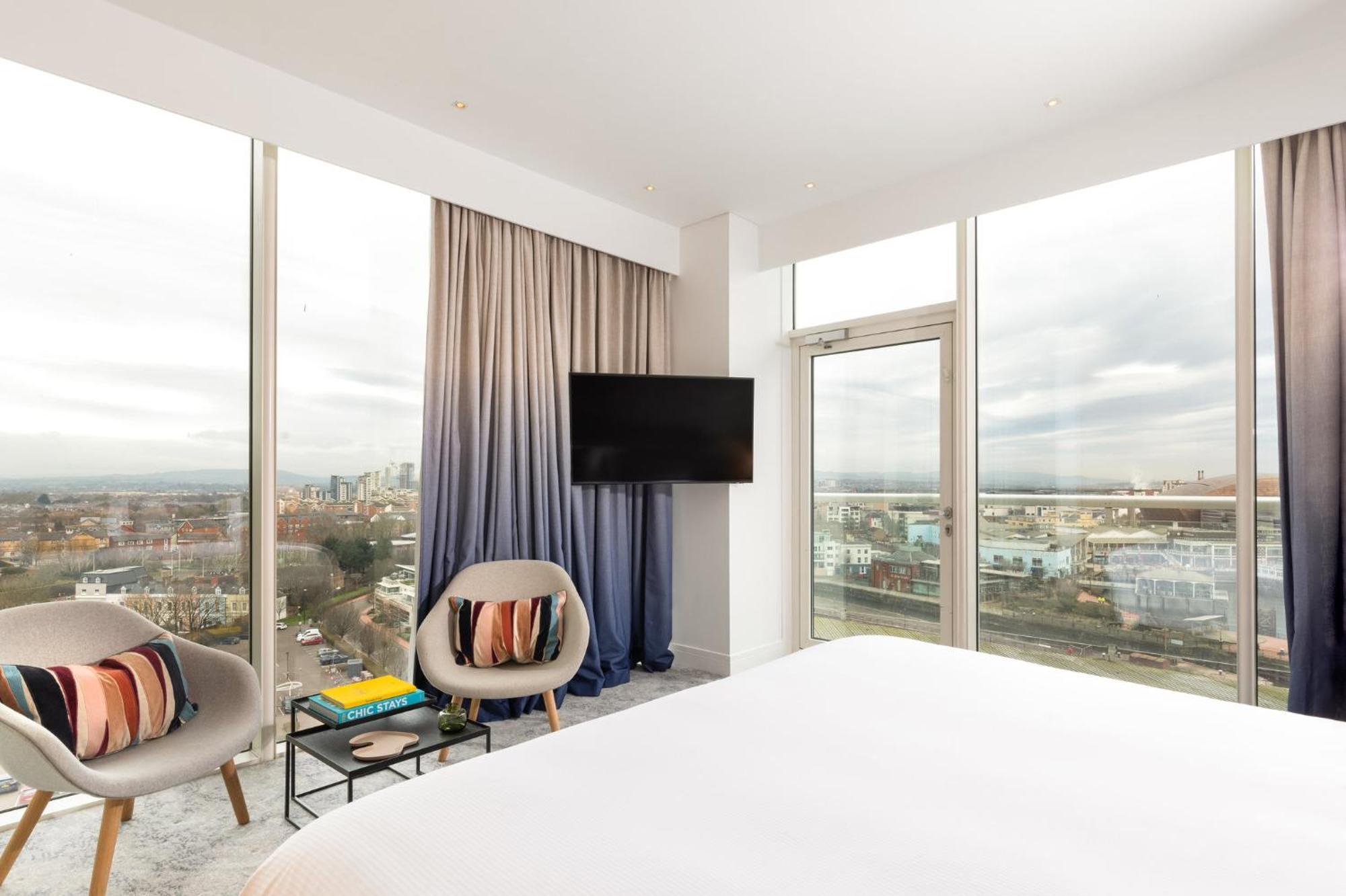 فندق فوكو سانت دايفيدز كارديف المظهر الخارجي الصورة The photo shows a modern hotel room featuring large windows with a view of the outside. The room is well-lit and has a clean, minimalist design. There are two stylish chairs with cushions, a small table, and a television mounted on the wall. The bed 