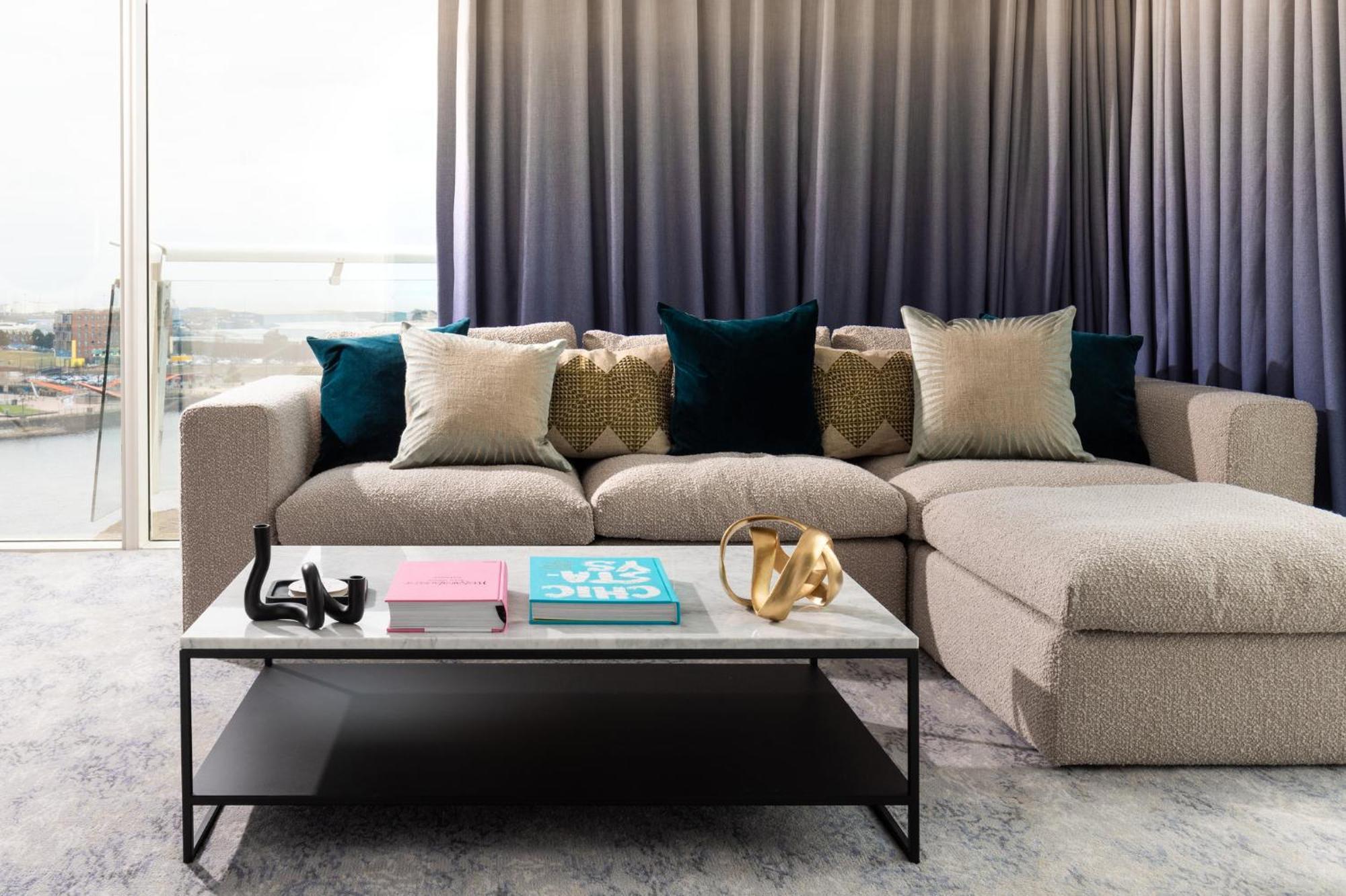 فندق فوكو سانت دايفيدز كارديف المظهر الخارجي الصورة The photo features a cozy living area with a spacious, L-shaped sofa upholstered in a light, textured fabric. The sofa is adorned with several decorative pillows in various colors, including soft beige and deep teal. In front of the sofa, there is a 
