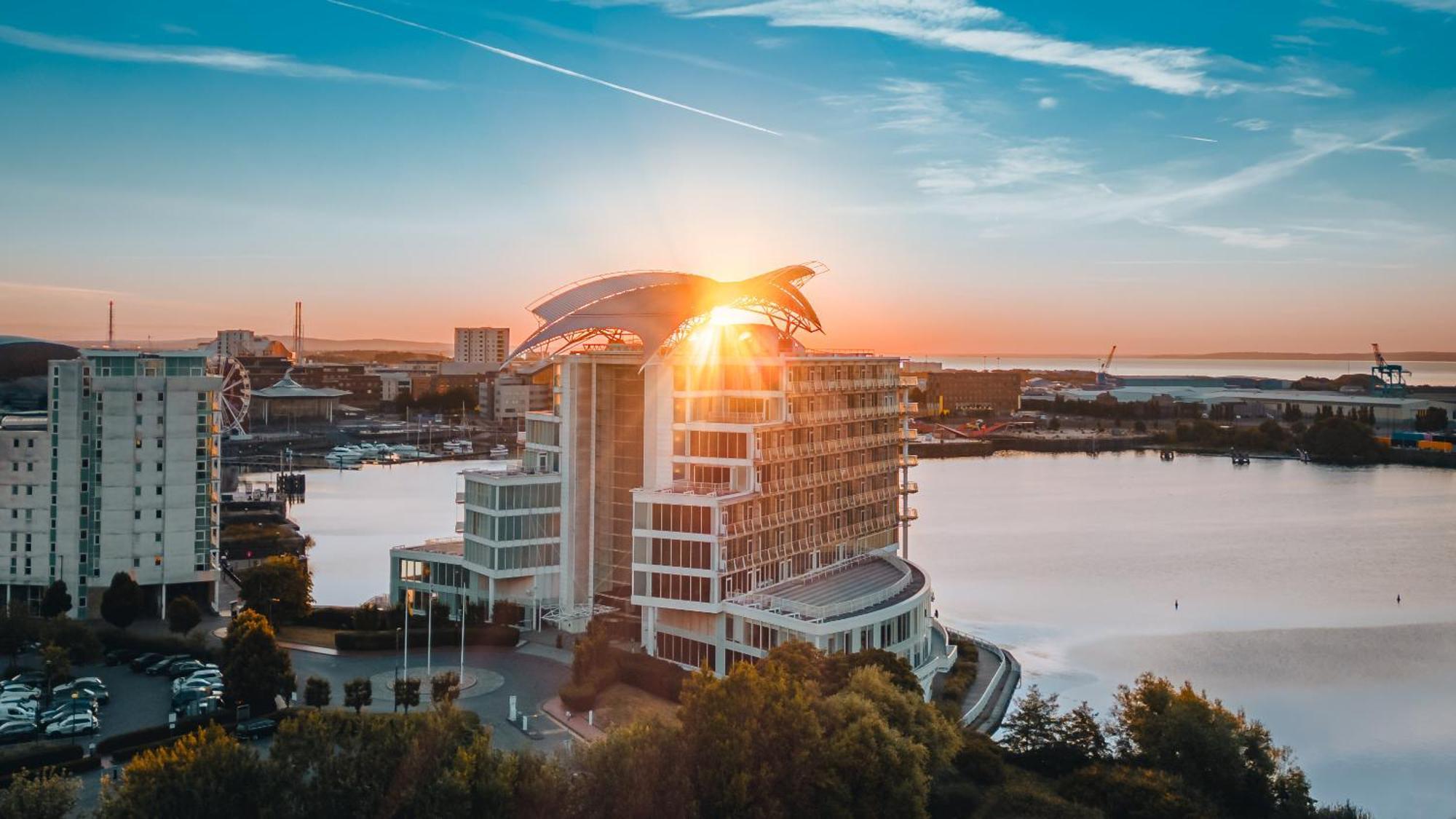 فندق فوكو سانت دايفيدز كارديف المظهر الخارجي الصورة The photo showcases a modern hotel building situated near a body of water, likely a bay or lake. The building is designed with a contemporary style, featuring large windows and a distinctive roofline that resembles a wave or bird. The sun is setting 