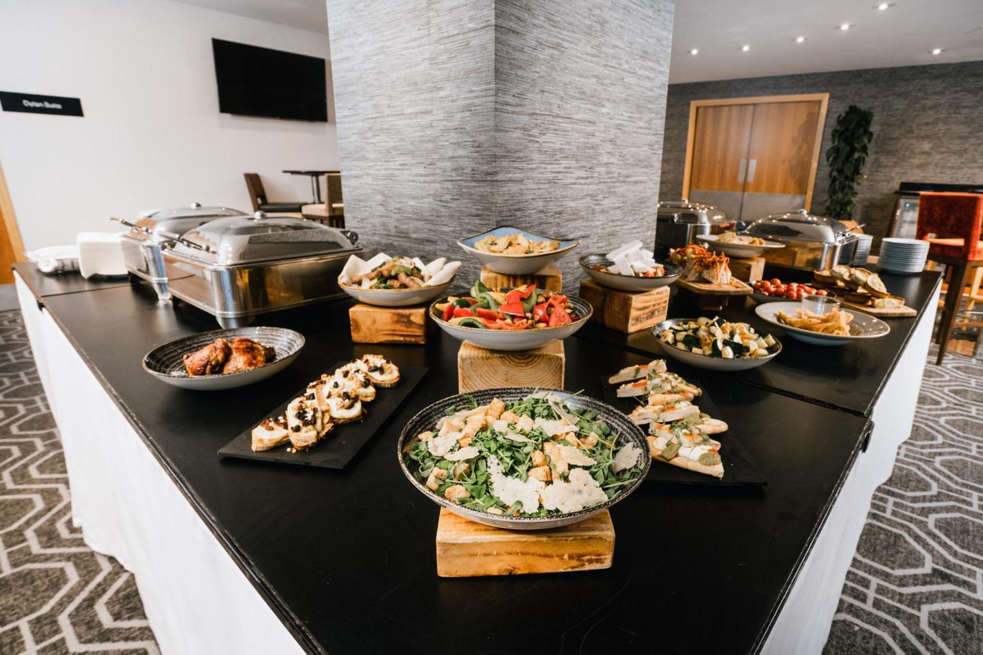 فندق فوكو سانت دايفيدز كارديف المظهر الخارجي الصورة The photo shows a buffet setup featuring a variety of food items displayed on a large table. There are multiple serving dishes with different types of food, including salads, appetizers, and possibly main dishes. The items are presented on both woode
